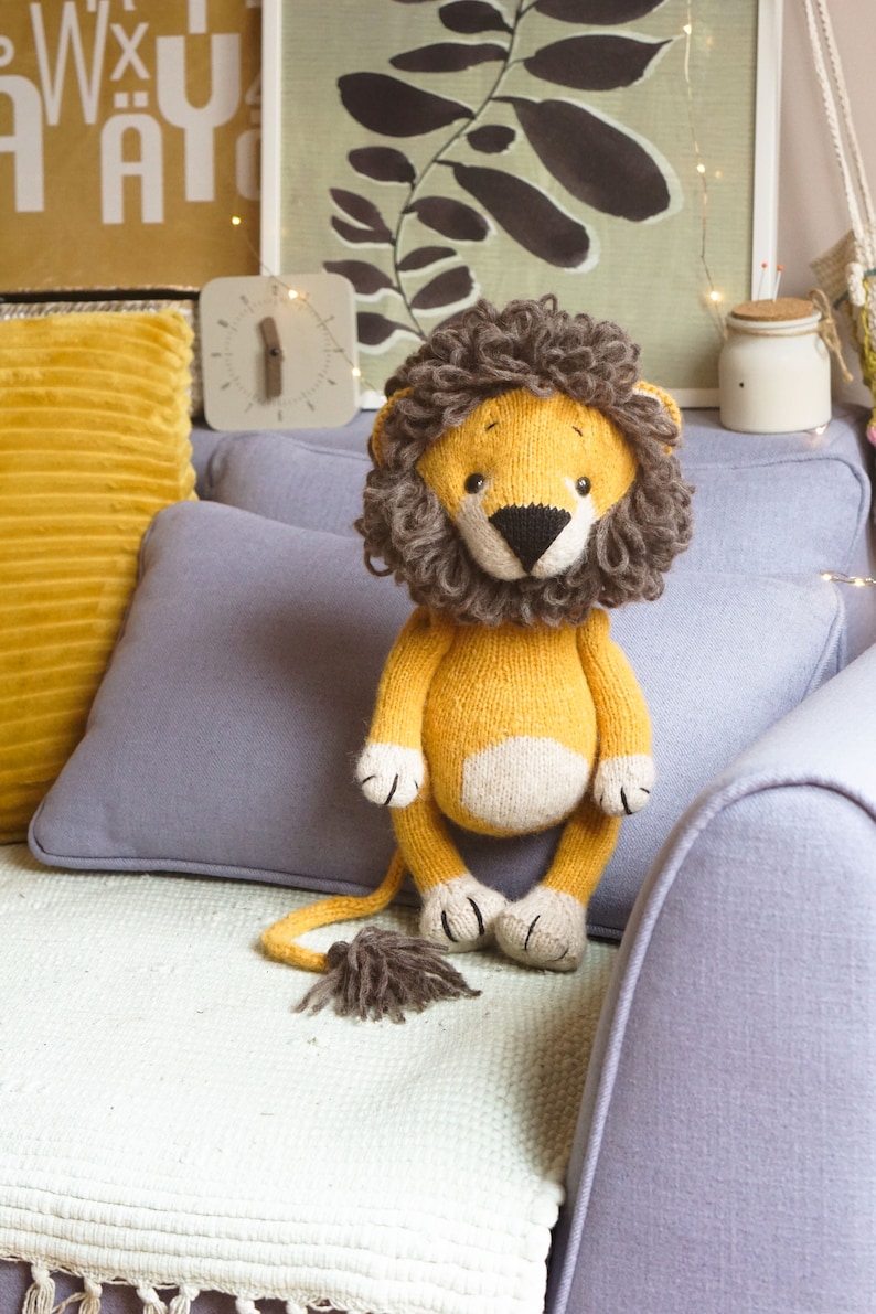 May include: A knitted lion toy with a brown mane, yellow body, and white belly. The toy is sitting on a light purple couch with a white blanket.
