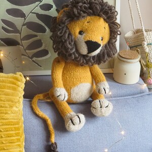 May include: A knitted lion toy with a brown mane, yellow body, and white paws. The toy is sitting on a blue couch with a yellow pillow and string lights.