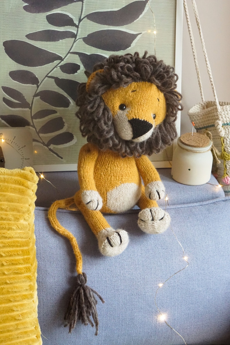 May include: A knitted lion toy with a brown mane, yellow body, and white paws. The toy is sitting on a blue couch with a yellow pillow and string lights.