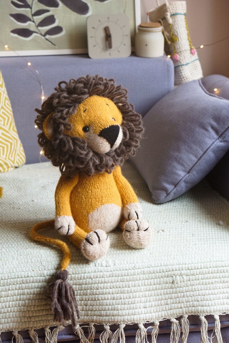 May include: A knitted lion toy with a brown mane, yellow body, and white paws. The toy is sitting on a light green blanket with a fringe.