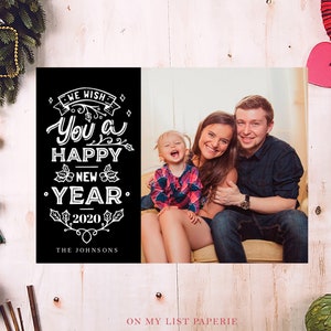 K&ouml;nnte beinhalten: Eine schwarz-wei&szlig;e Neujahrskarte mit dem Text "We wish you a Happy New Year 2020" und einem Familienportr&auml;t. Die Karte ist von Weihnachtsdekorationen umgeben, darunter ein Kranz, Ornamente und Zimtstangen.