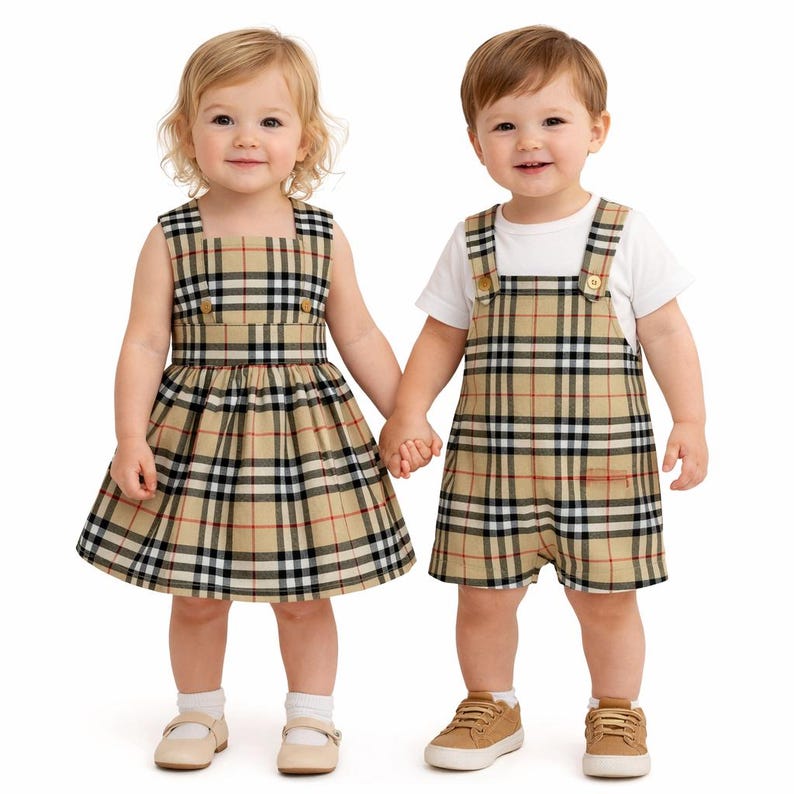 Pode incluir: Duas crian&ccedil;as de m&atilde;os dadas, vestindo roupas xadrez bege combinando. Uma usa um vestido sem mangas, a outra um macac&atilde;o sobre uma camiseta branca. Ambas as roupas apresentam um padr&atilde;o xadrez cl&aacute;ssico com detalhes em vermelho e preto.