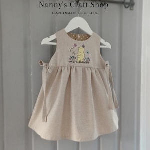 Op de afbeelding: Een beige mouwloze jurk met een geborduurde gele beer omringd door kleurrijke bloemen en vlinders. De jurk heeft striksluitingen aan de zijkanten en een voering met stippen. De tekst Nanny's Craft Shop HANDMADE CLOTHES is zichtbaar.