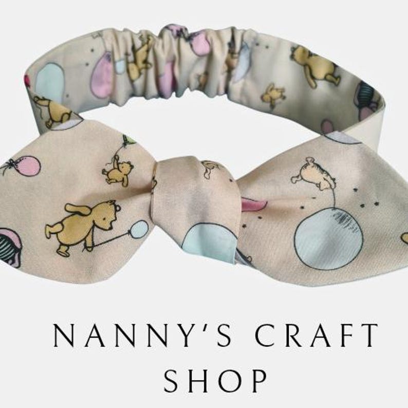Pode incluir: Uma faixa de cabelo bege com um la&ccedil;o com um desenho de urso de desenho animado. A faixa de cabelo tem uma se&ccedil;&atilde;o el&aacute;stica e o texto "NANNY'S CRAFT SHOP" em preto.