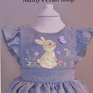 May include: A light blue dress with a bunny and butterfly embroidery. The dress features a round neckline, ruffled sleeves, and a gathered skirt. The text "Nanny's Craft Shop" is at the top of the image.