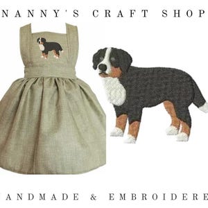 Op de afbeelding: Een saliegroene pinafore-jurk met een geborduurde Berner Sennenhond op de borst. De tekst NANNY'S CRAFT SHOP staat bovenaan en HANDMADE & EMBROIDERED onderaan. Een grotere versie van het hondenborduurwerk wordt apart weergegeven.