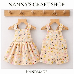 Pode incluir: Dois conjuntos de beb&eacute; feitos &agrave; m&atilde;o: um vestido e um macac&atilde;o. Ambos s&atilde;o bege com um padr&atilde;o repetido de personagens de desenhos animados e bal&otilde;es em amarelo, rosa e azul. Os conjuntos est&atilde;o pendurados em cabides de madeira com a parte superior em forma de urso, expostos contra uma parede clara. O texto "Nanny's Craft Shop" e "Handmade" s&atilde;o vis&iacute;veis.