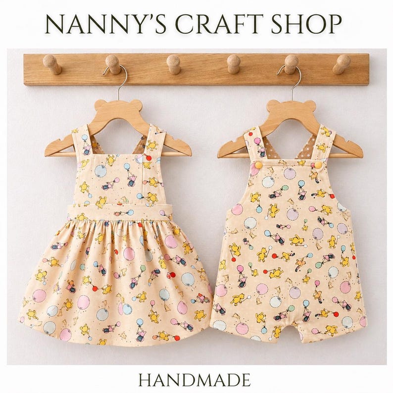 Pode incluir: Dois conjuntos de beb&eacute; feitos &agrave; m&atilde;o: um vestido e um macac&atilde;o. Ambos s&atilde;o bege com um padr&atilde;o repetido de personagens de desenhos animados e bal&otilde;es em amarelo, rosa e azul. Os conjuntos est&atilde;o pendurados em cabides de madeira com a parte superior em forma de urso, expostos contra uma parede clara. O texto "Nanny's Craft Shop" e "Handmade" s&atilde;o vis&iacute;veis.
