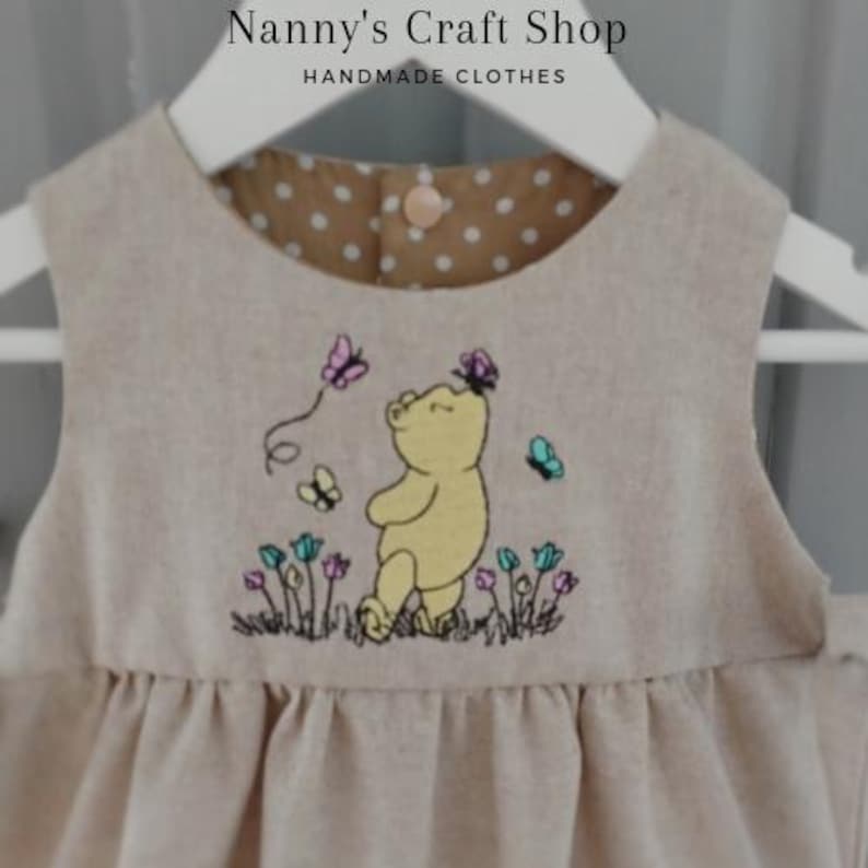 Op de afbeelding: Een beige mouwloze jurk met een geborduurde gele beer omringd door kleurrijke bloemen en vlinders. De binnenkant van de halslijn is gevoerd met bruine stof met witte stippen en heeft een enkele knoopsluiting aan de achterzijde.