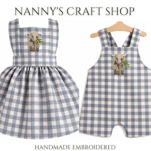 Pode incluir: Dois conjuntos infantis com padr&atilde;o xadrez azul e branco. Um &eacute; um vestido, o outro um macac&atilde;o. Ambos apresentam um bordado de vaca Highland e o texto "Nanny's Craft Shop". "Handmade Embroidered" tamb&eacute;m &eacute; vis&iacute;vel.