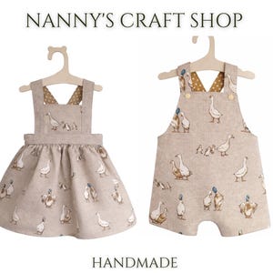 Pode incluir: Dois conjuntos infantis feitos &agrave; m&atilde;o em cabides. Um &eacute; um vestido, o outro &eacute; um macac&atilde;o. Ambos s&atilde;o bege com um padr&atilde;o repetido de gansos. O texto "Nanny's Craft Shop" e "Handmade" est&atilde;o na parte superior e inferior da imagem.