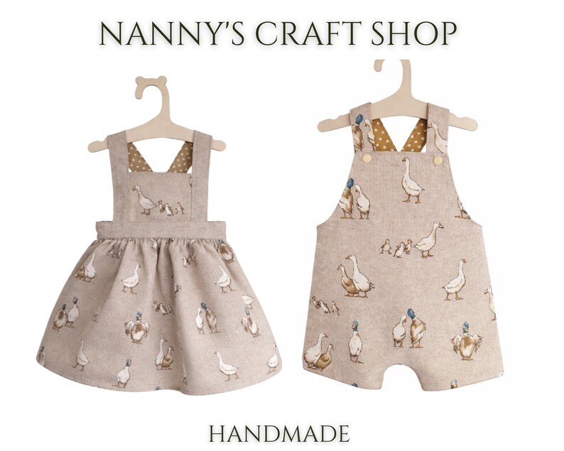 Pode incluir: Dois conjuntos infantis feitos &agrave; m&atilde;o em cabides. Um &eacute; um vestido, o outro &eacute; um macac&atilde;o. Ambos s&atilde;o bege com um padr&atilde;o repetido de gansos. O texto "Nanny's Craft Shop" e "Handmade" est&atilde;o na parte superior e inferior da imagem.