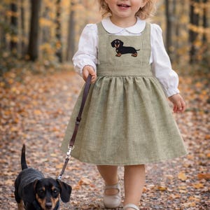 Pode incluir: Uma menina com um vestido verde com um aplique de dachshund passeia com um c&atilde;o dachshund com uma trela. O vestido &eacute; combinado com uma blusa branca e sapatos brancos. O c&atilde;o &eacute; preto e castanho. A cena acontece num caminho coberto de folhas ca&iacute;das.