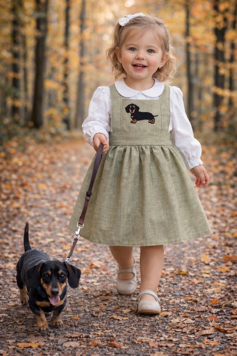 Pode incluir: Uma menina com um vestido verde com um aplique de dachshund passeia com um c&atilde;o dachshund com uma trela. O vestido &eacute; combinado com uma blusa branca e sapatos brancos. O c&atilde;o &eacute; preto e castanho. A cena acontece num caminho coberto de folhas ca&iacute;das.