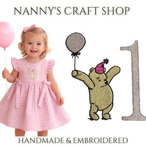 Op de afbeelding: Een roze jurk met vichy-ruitjes, ruches aan de mouwen en een strik, voorzien van een geborduurde beer met een feesthoedje die een ballon vasthoudt naast het cijfer 1. De tekst NANNY'S CRAFT SHOP en HANDMADE & EMBROIDERED staat op de afbeelding.