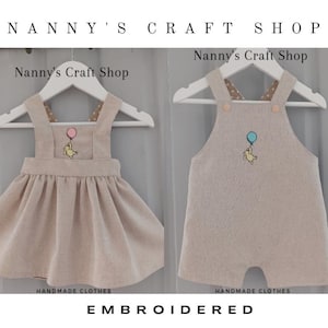 Pode incluir: Dois conjuntos de beb&eacute; bordados bege em cabides brancos. Um &eacute; um vestido com saia rodada, o outro &eacute; um macac&atilde;o. Ambos t&ecirc;m um desenho do Ursinho Pooh com um bal&atilde;o. O texto "Nanny's Craft Shop" e "Embroidered" s&atilde;o vis&iacute;veis.