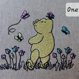 Op de afbeelding: Een borduurontwerp op lichtgrijze stof met een gele beer die tussen kleine blauwe en paarse bloemen staat. Drie vlinders in paarse en blauwe kleuren zijn rond de beer afgebeeld, waarvan er &eacute;&eacute;n op zijn neus rust. De tekst One is zichtbaar.