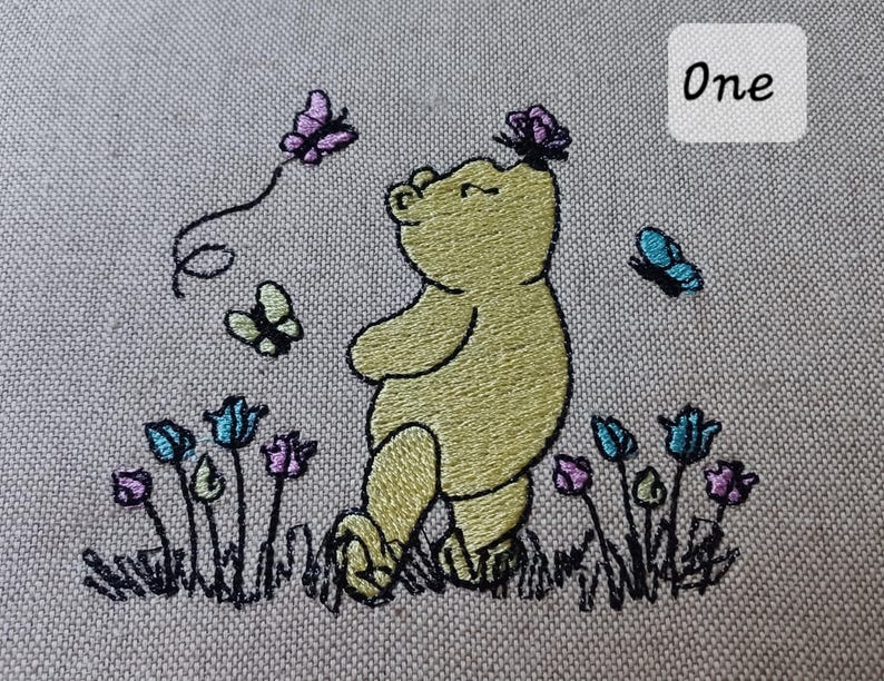 Op de afbeelding: Een borduurontwerp op lichtgrijze stof met een gele beer die tussen kleine blauwe en paarse bloemen staat. Drie vlinders in paarse en blauwe kleuren zijn rond de beer afgebeeld, waarvan er &eacute;&eacute;n op zijn neus rust. De tekst One is zichtbaar.