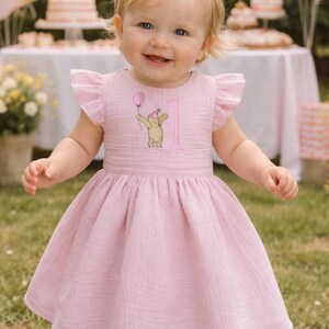 Pode incluir: Vestido rosa claro com mangas esvoa&ccedil;antes e uma faixa de cabelo a condizer. O vestido apresenta um desenho bordado de um urso segurando um bal&atilde;o e o n&uacute;mero 1. A crian&ccedil;a sorri, em p&eacute; na relva, com uma festa ao fundo.