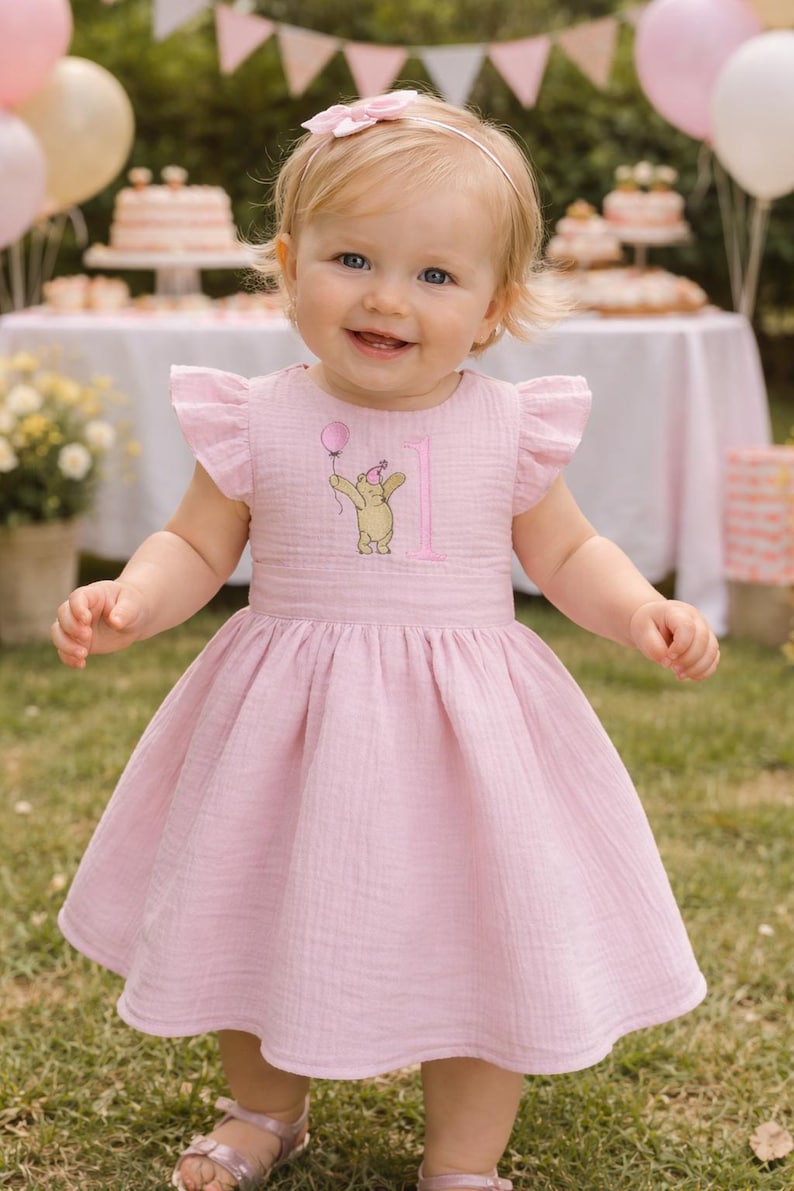 Pode incluir: Vestido rosa claro com mangas esvoa&ccedil;antes e uma faixa de cabelo a condizer. O vestido apresenta um desenho bordado de um urso segurando um bal&atilde;o e o n&uacute;mero 1. A crian&ccedil;a sorri, em p&eacute; na relva, com uma festa ao fundo.