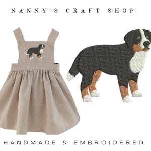 May include: A beige linen dress with a Bernese Mountain Dog embroidery. The dress has a square neckline and suspender straps. A separate embroidered Bernese Mountain Dog is also shown. The text "Nanny's Craft Shop" and "Handmade & Embroidered" are visible.