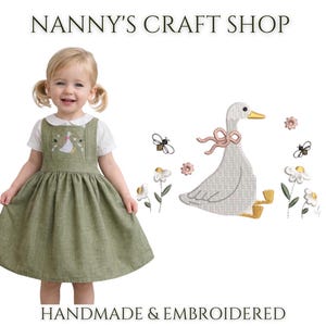 Op de afbeelding: Een saliegroene overgooier over een wit overhemd met kraag. De jurk is voorzien van een borduursel van een witte gans met een roze lint, omringd door kleine bloemen en bijen. De tekst NANNY'S CRAFT SHOP HANDMADE & EMBROIDERED is aanwezig.