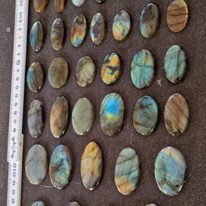 May include: Twenty-five oval-shaped labradorite stones with iridescent blue, green, and gold hues. The stones are arranged in five rows of five on a brown surface.
