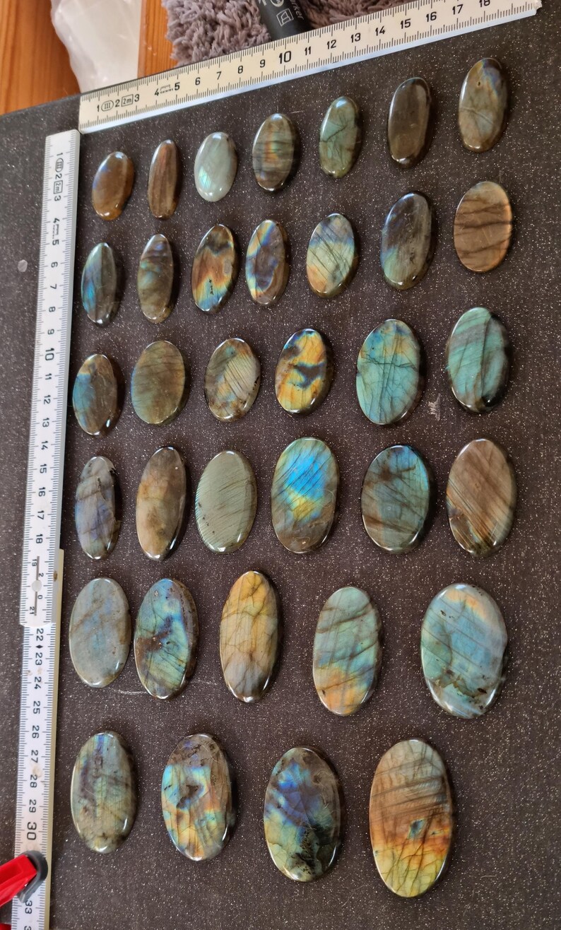 May include: Twenty-five oval-shaped labradorite stones with iridescent blue, green, and gold hues. The stones are arranged in five rows of five on a brown surface.