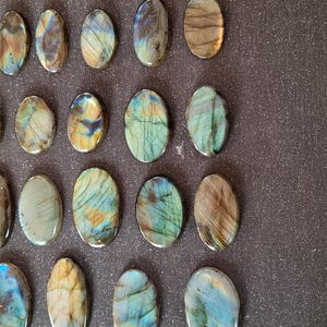 May include: A collection of 18 oval-shaped labradorite stones, each with a unique iridescent sheen. The stones range in color from dark gray to light blue, with hints of green and gold. The stones are arranged in rows on a dark gray surface.