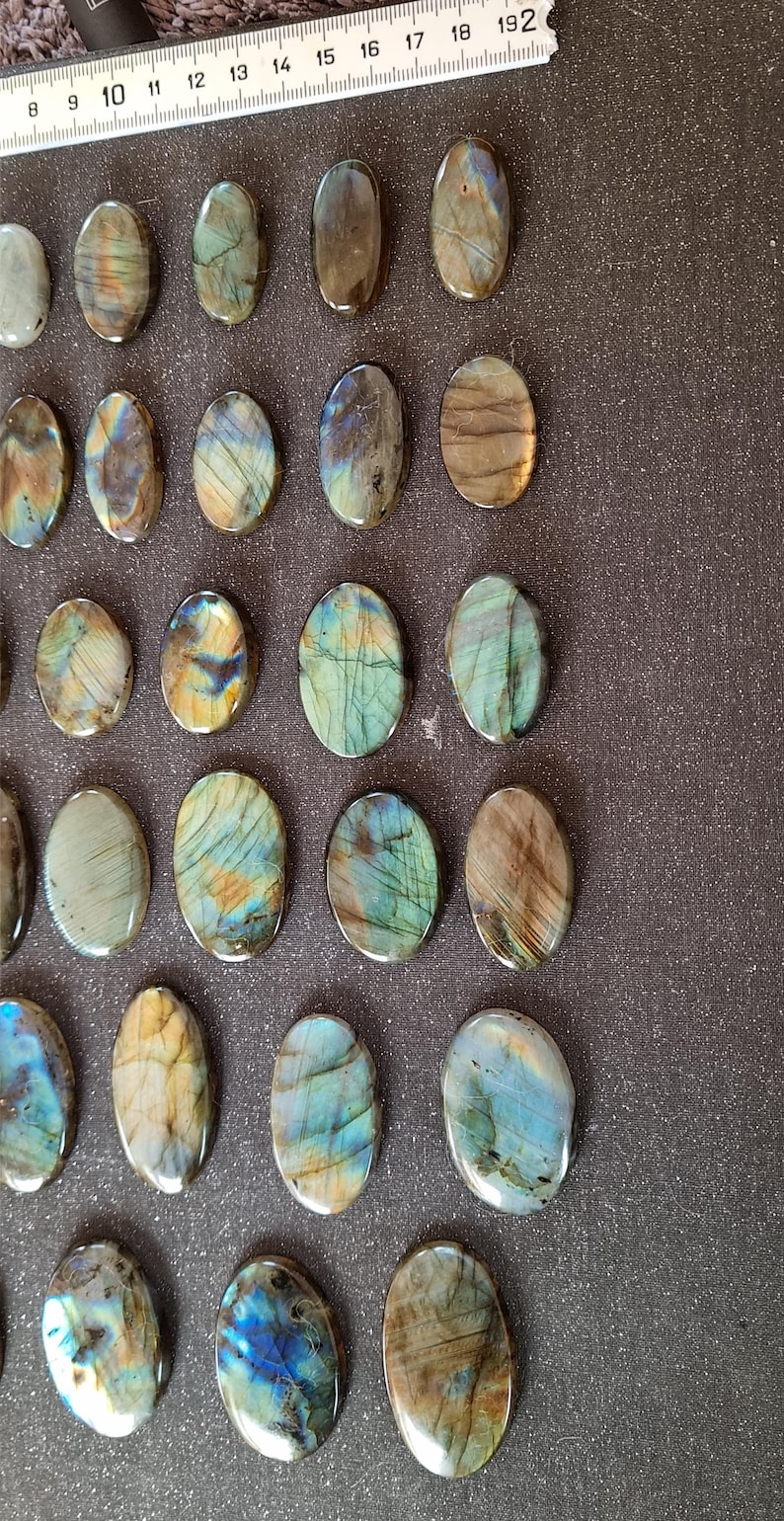 May include: A collection of 18 oval-shaped labradorite stones, each with a unique iridescent sheen. The stones range in color from dark gray to light blue, with hints of green and gold. The stones are arranged in rows on a dark gray surface.