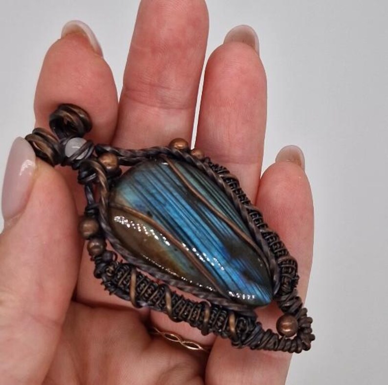 K&ouml;nnte beinhalten: Ein handgefertigter Anh&auml;nger mit einem blauen und gr&uuml;nen Labradorit-Edelstein. Der Stein ist in eine Kupferdrahtwicklung mit aufwendigen Details eingefasst. Der Anh&auml;nger hat eine Tropfenform und ist an einem Kupferdraht aufgeh&auml;ngt. Der Edelstein hat einen schillernden Glanz.