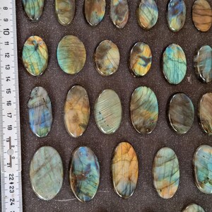 May include: A collection of 28 oval-shaped labradorite stones, each with a unique iridescent pattern of blue, green, and gold. The stones are arranged in rows on a dark brown surface.