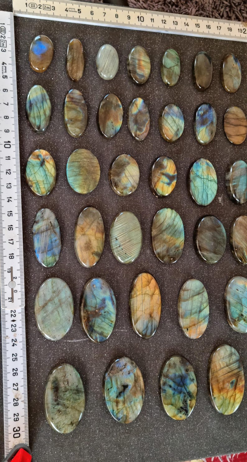 May include: A collection of 28 oval-shaped labradorite stones, each with a unique iridescent pattern of blue, green, and gold. The stones are arranged in rows on a dark brown surface.