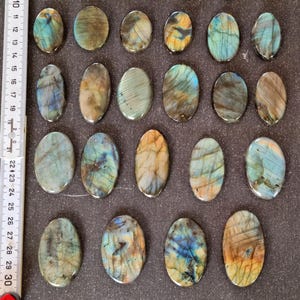 May include: Twenty oval-shaped labradorite stones with a variety of blue, green, and gold colors. The stones are arranged in rows on a dark brown surface.