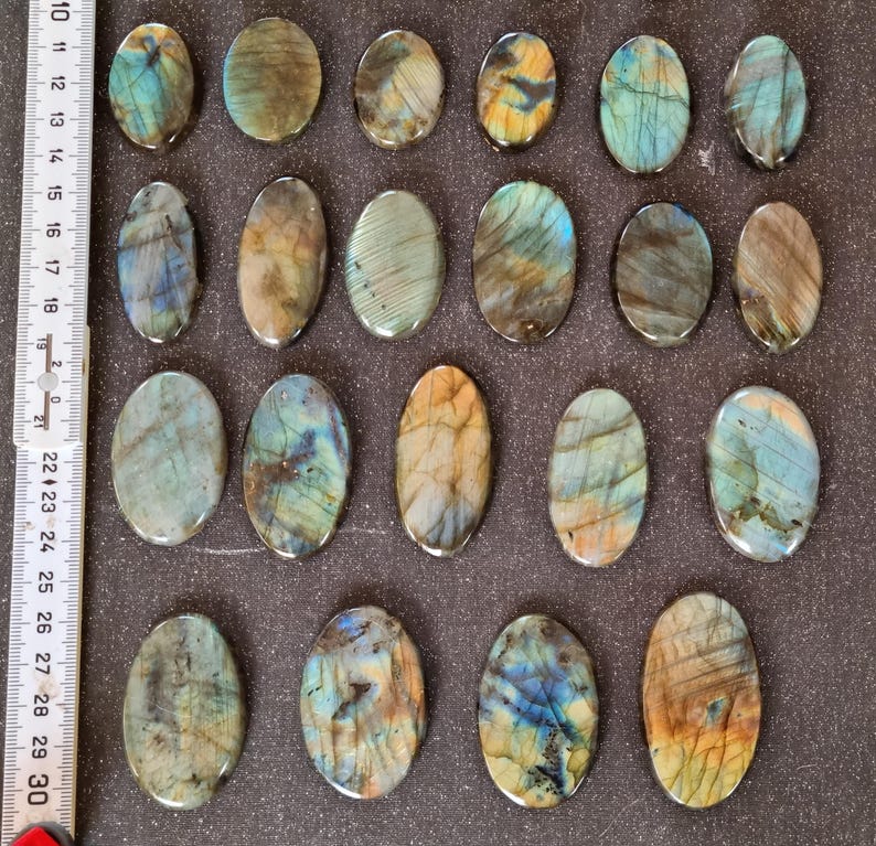 May include: Twenty oval-shaped labradorite stones with a variety of blue, green, and gold colors. The stones are arranged in rows on a dark brown surface.
