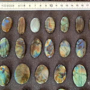 May include: Twenty oval-shaped labradorite stones, each with a unique iridescent sheen, ranging from green to blue to gold. The stones are arranged in three rows on a gray surface.