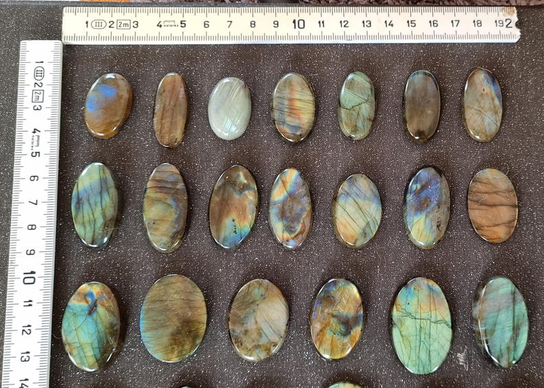 May include: Twenty oval-shaped labradorite stones, each with a unique iridescent sheen, ranging from green to blue to gold. The stones are arranged in three rows on a gray surface.