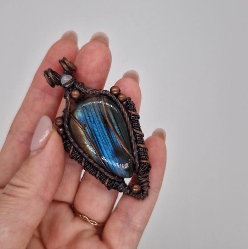 K&ouml;nnte beinhalten: Handgefertigter Anh&auml;nger mit einem blauen und goldenen Labradoritstein, kunstvoll in Kupferdraht gewickelt. Der Anh&auml;nger hat eine Tropfenform und ist mit kleinen Kupferperlen und einem kleinen wei&szlig;en Stein verziert.