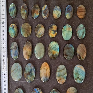May include: Twenty-five oval-shaped labradorite stones, ranging in color from dark gray to brown with iridescent blue and green hues. The stones are arranged in five rows of five on a dark gray surface.
