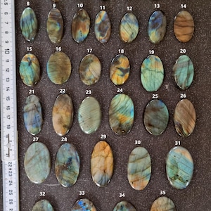 May include: Thirty-five oval-shaped labradorite gemstones, each with a unique iridescent pattern of blue, green, gold, and brown. The stones are arranged in rows on a dark gray surface, numbered 1 through 35.