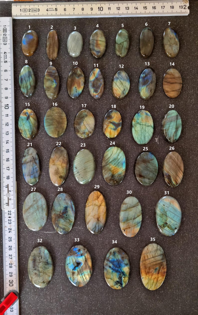 May include: Thirty-five oval-shaped labradorite gemstones, each with a unique iridescent pattern of blue, green, gold, and brown. The stones are arranged in rows on a dark gray surface, numbered 1 through 35.
