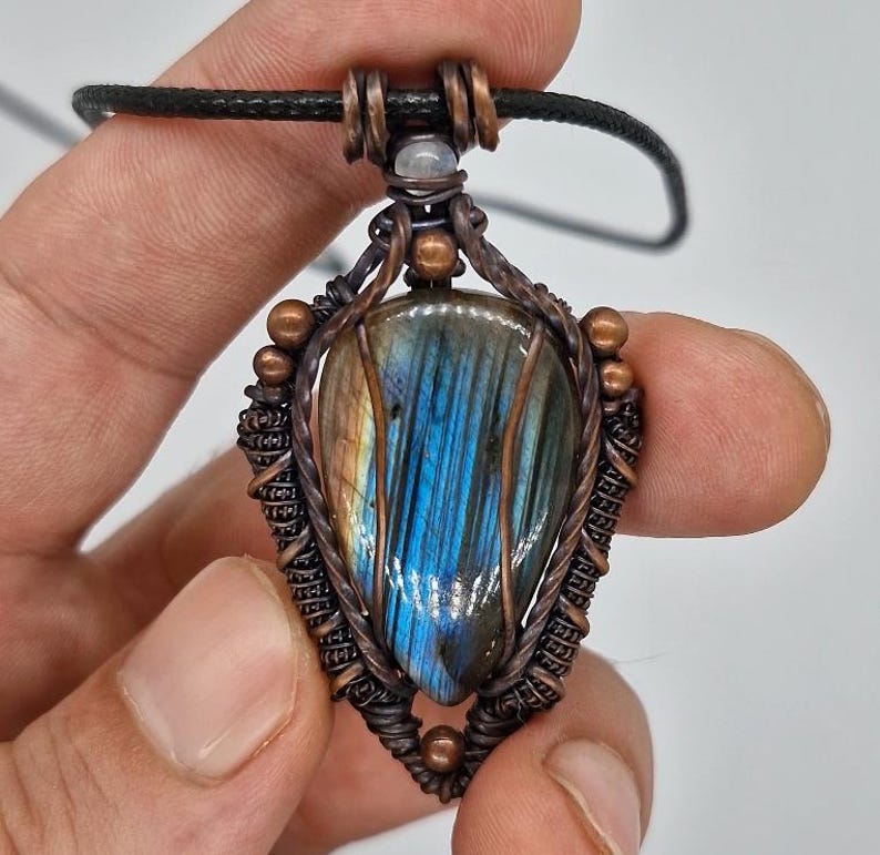 K&ouml;nnte beinhalten: Ein handgefertigter Anh&auml;nger mit einem tropfenf&ouml;rmigen Labradoritstein mit blau-goldenen Irisierungen. Der Stein ist in aufwendiger Kupferdrahtarbeit gefasst und an einer schwarzen Kordel befestigt. Der Anh&auml;nger wird in einer Hand gehalten.