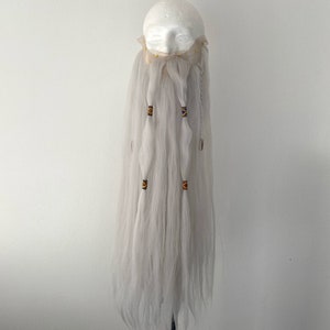 May include: A long, gray beard prop with multiple braided sections. Each braid is adorned with small, decorative beads in shades of brown, yellow, and orange. The beard is attached to a foam head, suggesting a costume or theatrical accessory.