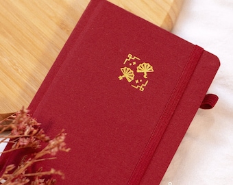 May include: A burgundy notebook with a gold elastic closure and a gold design on the front. The design features a stylized fan with stars and a crescent moon. The notebook has the text "poyuro.co" printed in gold on the bottom right corner.