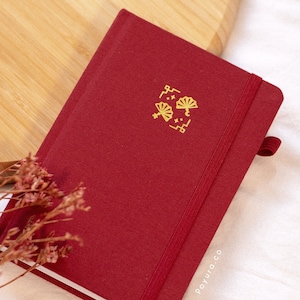 May include: A burgundy notebook with a gold elastic closure and a gold design on the front. The design features a stylised fan with stars and a crescent moon. The notebook has the text "poyuro.co" printed in gold on the bottom right corner.