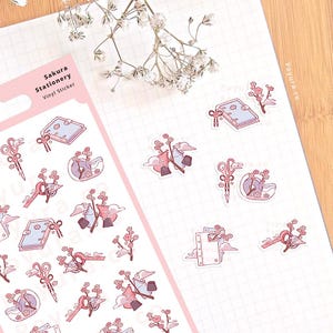 Op de afbeelding: Een vel Sakura Stationery vinyl stickers. De stickers tonen roze en witte kersenbloesems, scharen en andere Japanse ontwerpen. De sticker heeft de tekst "Sakura Stationery Vinyl Sticker".