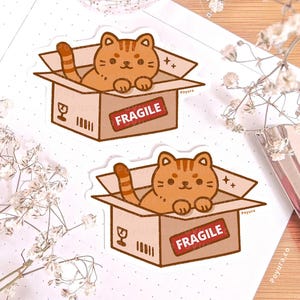 Op de afbeelding: Twee stickers met een oranje tabby kat die uit een kartonnen doos kijkt. De dozen hebben een rood "FRAGILE" label en verzendsymbolen. De stickers liggen op een wit oppervlak met bloemmotieven. De naam van de kunstenaar, "Poyura", is zichtbaar.