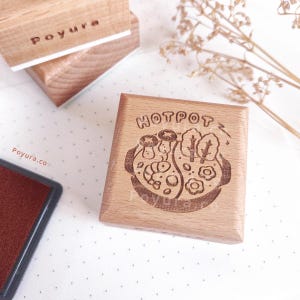 Könnte beinhalten: Holzstempel mit dem Wort "HOTPOT" und einer Cartoon-Illustration eines Hot Pots mit Pilzen, Gemüse und Eiern. Der Stempel ist aus hellem Holz gefertigt. Der Markenname "Poyura" ist auf dem Stempel und dem Stempelkissen zu sehen.