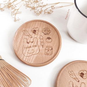 May include: Wooden coaster with engraved food illustrations, including a bag with "THANK YOU" text, sushi, and a milk carton. The coaster is round and made of light-colored wood. A white mug is visible in the background.