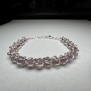 May include: A silver bracelet featuring a chainmail design, with small, round, light purple amethyst beads. The bracelet is made of silver-colored metal and has a clasp closure. The jewelry is displayed on a white surface.